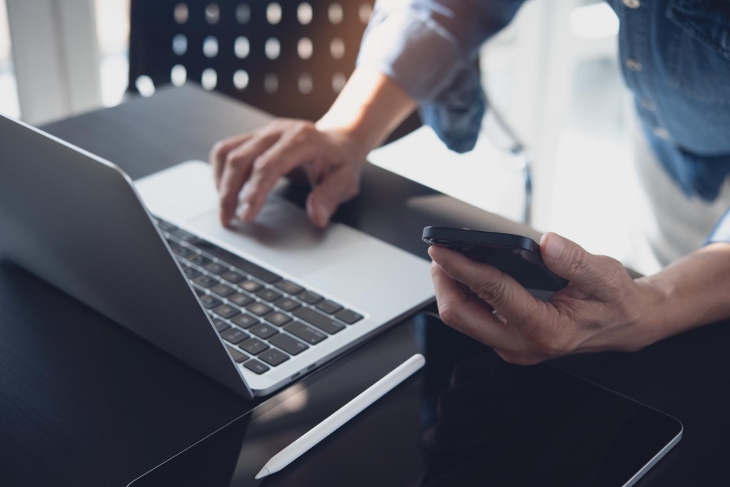 Close up of man using smart phone and working on laptop computer on wooden table with blurred background